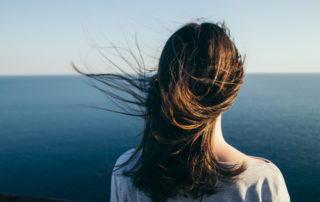 Woman with dark hair stands on a top cliff over blue sea view while wind.