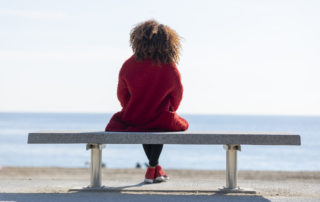 Rear view of young curly woman wearing red denim jacket sitting on a bench while looking away to horizon over sea