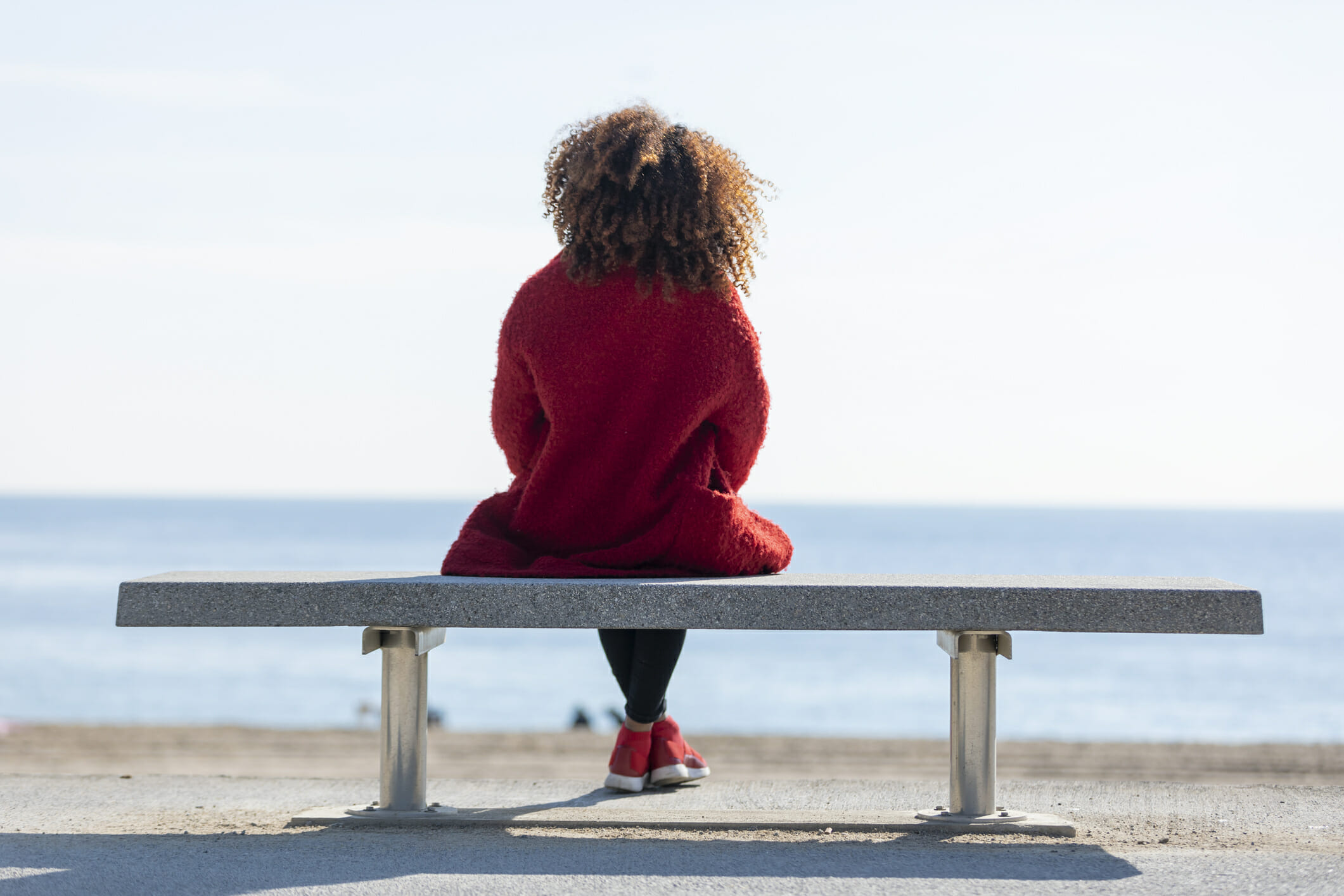 Rear view of young curly woman wearing red denim jacket sitting on a bench while looking away to horizon over sea