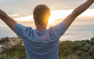 Cheerful young man arms outstretched at sunset, coastline on the background.