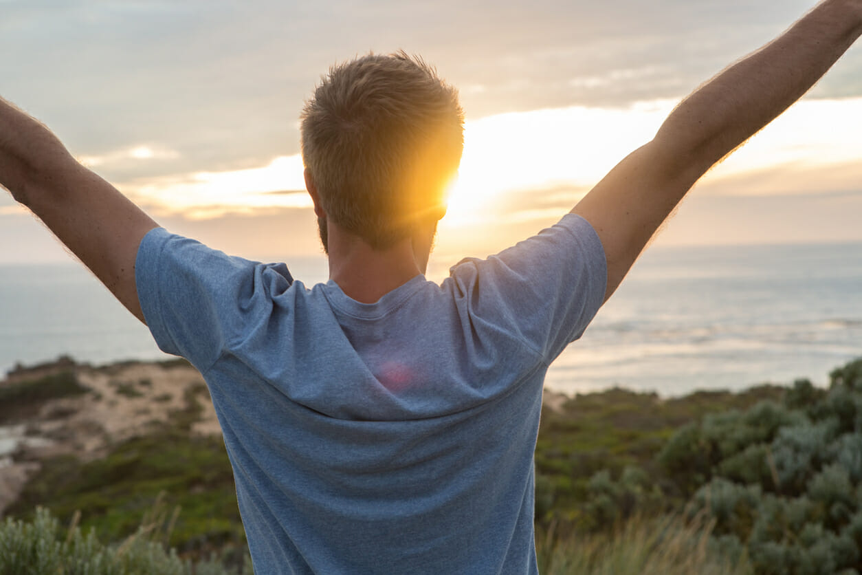 Cheerful young man arms outstretched at sunset, coastline on the background.